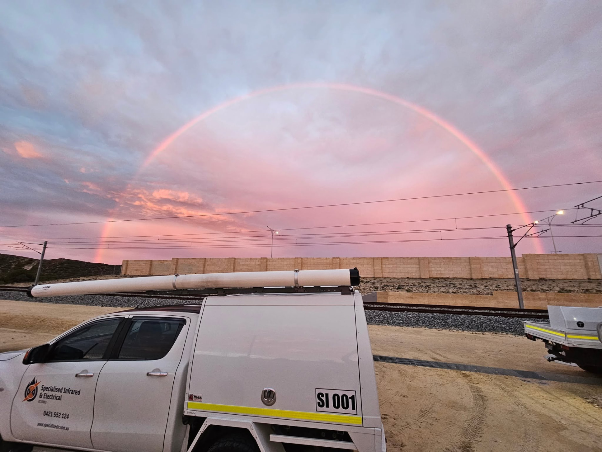 Specialised Infrared & Electrical service ute parked at a rail corridor under a rainbow at sunset